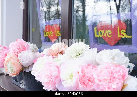 Weiß-rosa Pfingstrosen auf der Fensterbank vor dem Hintergrund der Inschrift Love is. Stockfoto