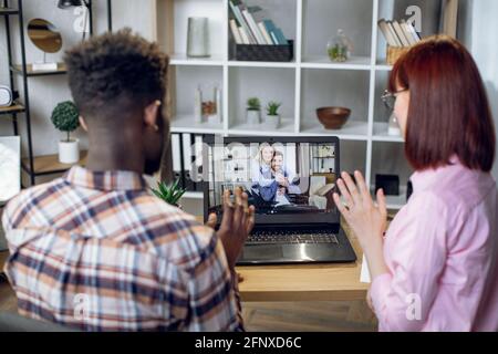Rückansicht eines multirassischen Paares, das am Tisch sitzt und während eines Videoanrufs auf einem modernen Laptop mit kaukasischen Freunden winkt. Glückliche, vielfältige Menschen, die während der Quarantäne aus der Ferne sprechen. Stockfoto
