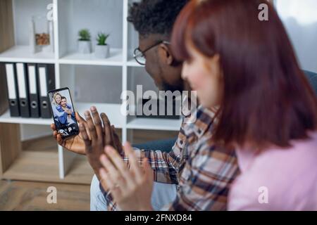 Rückansicht eines multiethnischen Paares, das während eines Videogesprächs mit Freunden auf dem Smartphone die Hände winkt. Menschen, die moderne Technologie nutzen, während sie zu Hause bleiben. Stockfoto