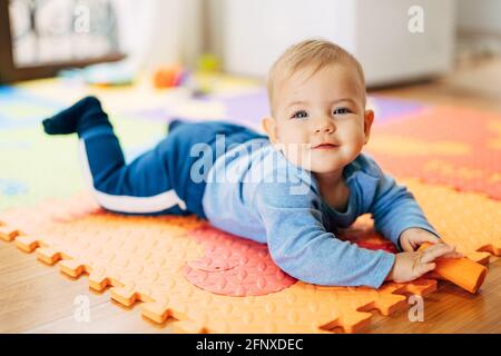 Lächelnder kleiner Junge in einem blauen Anzug mit einem Karotte in den Händen liegt mit seinem Bauch auf einem Farbiger Teppich auf dem Boden vor dem Hintergrund der Stockfoto