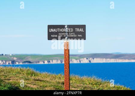 Nicht autorisiertes Wanderzeichen, das Wanderer vor gefährlichen Klippen warnt. Verschwommene malerische Küstenlinie mit dramatischen Sandsteinklippen der Drakes Bay in Backgrou Stockfoto