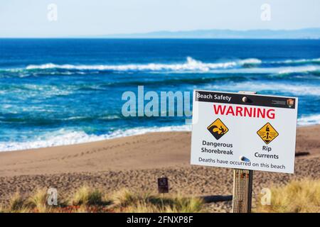 Das Warnschild am Strand informiert Besucher über gefährliche Küstenabrissströme und reißende Strömungen. Verschwommene Meereswellen und Sandstrand im Hintergrund. - Point Reye Stockfoto