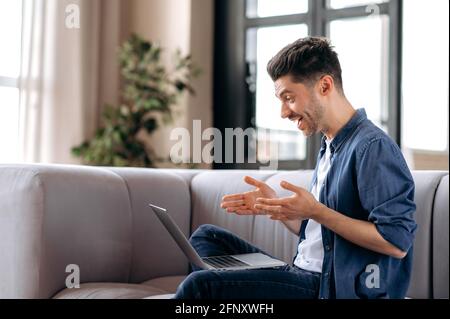 Videokommunikation, Online-Konferenz. Der junge Erwachsene kaukasische Mann, Freiberufler oder Student, in Jeanshemd, sitzt auf einem Sofa im Wohnzimmer und kommuniziert mit seinen Kollegen per Videoanruf über einen Laptop Stockfoto