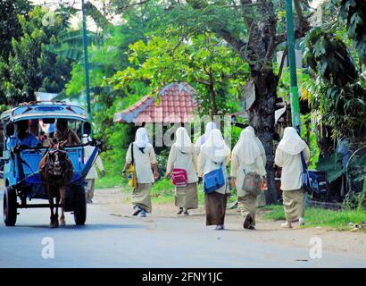 Eine Gruppe Studentinnen, die am Straßenrand zur Schule gehen, während sich im Straßenzentrum in Lombok, Indonesien, ein Pferdewagen bewegt. Stockfoto