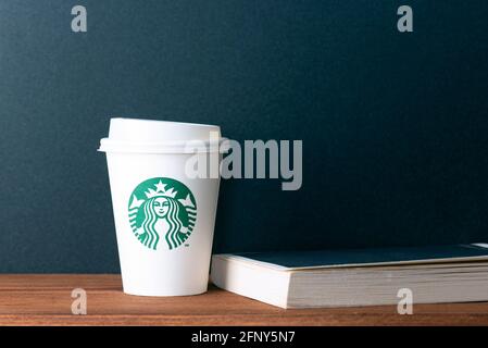 BERLIN - MAI 19: Weißer Becher aus Papier mit Starbucks-Logo und Buch auf Holztisch in Berlin, Mai 19. 2021 in Deutschland. Stockfoto