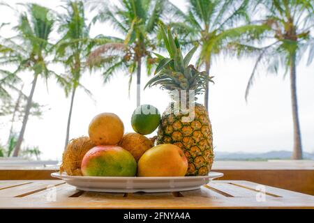 Ein Teller mit exotischen Früchten auf einem Holztisch mit Palmen im Hintergrund. Stockfoto