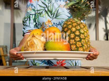 Frau trägt ein lebendiges Kleid mit einem großen Tablett Ganze tropische Früchte über einem Holztisch Stockfoto