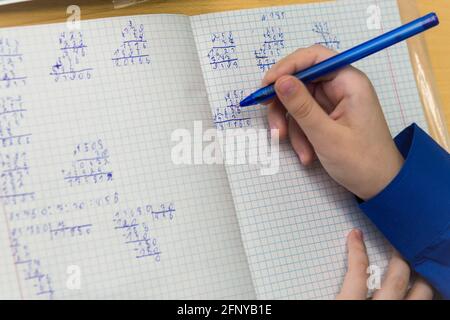 Eine Mädchenhand mit Kugelschreiber schreibt in schlechter Handschrift in Nahaufnahme. Der Studierende schließt die Aufgabe ab. Ein Student löst an einem Schreibtisch in der Schule Beispiele in Mathematik. Warmes, weiches Tageslicht. Stockfoto