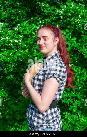 Das Mädchen hält einen niedlichen Ingwerkaninchen auf Sommer Natur im Park im Freien auf dem Hintergrund eines blühenden Baumes. Stockfoto