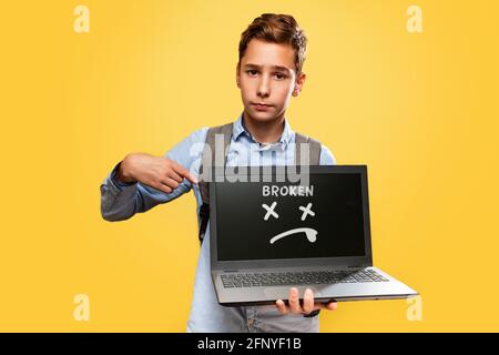 Konzept der modernen Technologien und Bildung. Ein verärgerter kaukasischer Teenager hält einen offenen Laptop und zeigt mit dem Finger darauf. Gelbe Rückseite Stockfoto