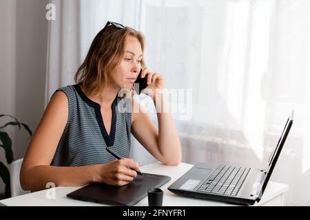 Remote-Arbeit. Eine Frau arbeitet zu Hause auf einem grafischen Tablet und spricht am Telefon. Das Konzept von Quarantäne, Freiberufung und Selbstisolation. Stockfoto