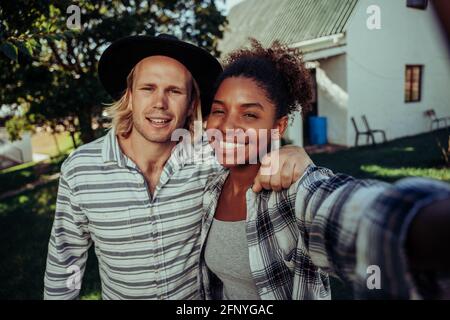 Gemischtes Rassenpaar lächelt beim Selfie mit stehendem Mobilfunkgerät Vor dem weißen Farmhaus Stockfoto