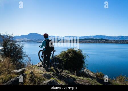 Eine Radfahrerin, die mit ihrem Fahrrad steht und die Aussicht auf den Lake Wanaka, South Island, genießt Stockfoto