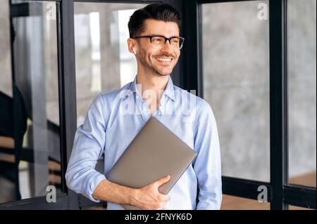 Porträt eines gutaussehenden, selbstbewussten, erfolgreichen kaukasischen Geschäftsmannes oder Büroangestellten, der Hemd und Brille trägt, mit einem Laptop im Büro steht, zur Seite schaut und freundlich lächelt Stockfoto