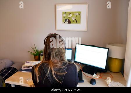 Rückansicht einer gesichtslosen jungen Frau, die während der COVID-19-Pandemie in Großbritannien auf digitalen Bildschirmen mit Kopfhörern zu Hause arbeitet KATHY DEWITT Stockfoto