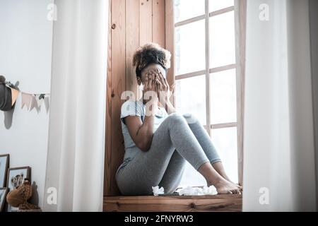 Frau bedeckt das Gesicht mit Händen, die auf dem Fensterbrett sitzen Stockfoto