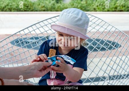 Mama füttert Baby im Park. Ein gesunder Snack. Ein Mädchen sitzt auf einem Straßenstuhl und hält einen Snack. Sie lächelt und ist glücklich Stockfoto