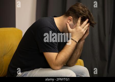 Der unrasierte Kerl sitzt auf dem Sofa und packte seinen Kopf in die Hände An Depressionen leiden Stockfoto