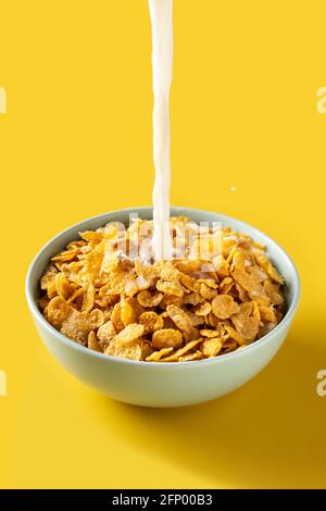 Milch in eine Schüssel mit Maisflocken auf gelbem Hintergrund gießen Stockfoto