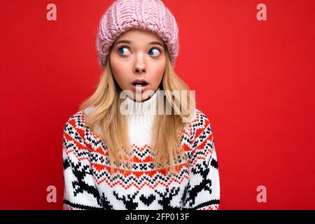 Foto von schönen schockiert junge blonde Frau isoliert über roten Hintergrund Wand tragen Winterpullover und rosa Hut Blick zur Seite Stockfoto
