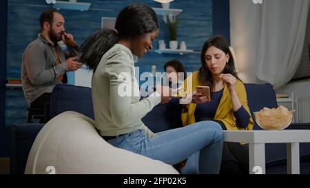 Mixed-Rennen Freunde sitzen auf Sofa-Bestände mobile in Händen Surfen im Internet, Online-Videos während einer entspannten Party zu sehen. Eine Gruppe multiethnischer Menschen, die gemeinsam im Wohnzimmer herumhängen Stockfoto