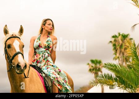 Porträt von jungen schönen Brünette Frau trägt Blumen Kleid Reiten braunes Pferd im Sommer bewölkten Tag. Stockfoto