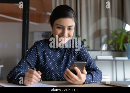 Lächelnde Inderin nimmt sich Notizen mit dem Smartphone Stockfoto
