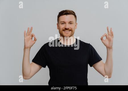 Junger Mann mit positivem Bart, der seine Zustimmung zeigte, während er eine gute Geste zeigte Mit beiden Händen Stockfoto