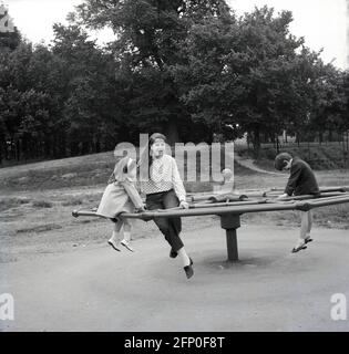 1960, historisch, draußen auf einem Spielplatz in einem öffentlichen Park, drei Kinder spielen auf einem Metallrad, das sich beim Drücken um eine zentrale Achse drehte, England, Großbritannien. Dieser riesige Metallkreis, der sich etwa einen Fuß vom Boden entfernt befand, war in dieser Zeit, manchmal auch als der „große Spinner“ bekannt, eine beliebte Fahrt auf öffentlichen Park-Spielplätzen. Stockfoto