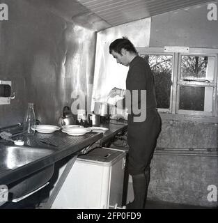 1960, historisch, in einer Sperrholzhütte oder Hütte, neben einem Edelstahlwaschbecken und einer Arbeitsplatte, ein Mann, der knickerbocker-Hosen trägt, um in einem Topf auf einem kleinen elektrischen Heizgerät Essen, vielleicht Suppe, zu erwärmen, England, Großbritannien. Könnte ein Tourenfahrer sein, wenn man den Hosenstil bedenkt, den er trägt. Diese in lange Socken gesteckte Hose mit Baggy-Kneed war ein beliebtes Kleidungsstück der Vorkriegszeit und blieb so bis in die 1950er und 1960er Jahre, vor allem in Großbritannien, bis sie von so genannten Rennstrumpfhosen oder Leggings in den Schatten gestellt wurden, obwohl die meisten Radfahrer nicht Rennen. Stockfoto