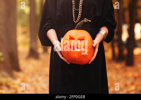 Junge Frau in schwarzem Umhang mit gruseligen Jack o Laterne Und auf den Halloween-Tag im Herbstwald wegschauen Stockfoto