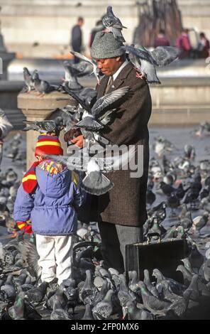 Archivmaterial aus den 1990er Jahren: Winterlicher Straßenszene Mann, der Taubenfutter von Hand füttert und neben seinem Aktenkoffer und seinem kleinen Kind Trafalgar Square London England Großbritannien Pflaster verwendet Stockfoto