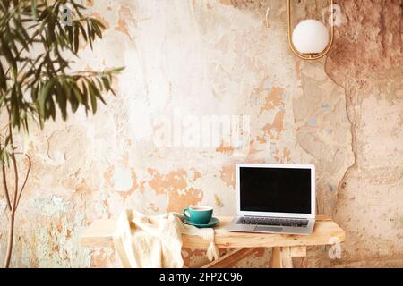 Retro-Tasse frischen Kaffee auf Strickmode fast modern platziert Netbook mit leerem Bildschirm auf Holztisch gegen grungigen Putz Wand im Kreativstudio Stockfoto