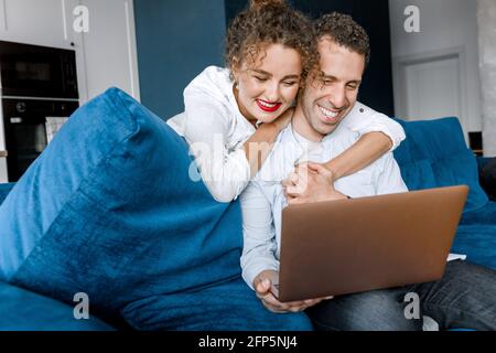 Gemeinsam das Leben beginnen. Inspiriertes Brautpaar kuschelt auf der Couch und plant den Kauf eines Laptops für ein neues Zuhause. Glücklich junges Paar in der Liebe wählen Stockfoto