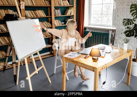 Glückliche junge schöne Studentin zeigt Daumen nach oben und genießt das Studium aus der Ferne. Fröhliche tausendjährige Frau, die auf den Computerbildschirm schaut, mit Stockfoto
