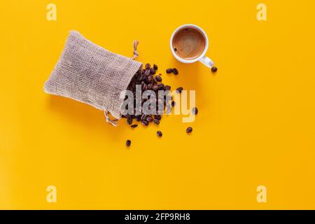 Es ist ein kleiner Sack offen und liegend, aus dem Kaffeebohnen fallen und streuen auf einem gelben Hintergrund von oben gesehen. Oben ist ein weißer Becher Stockfoto