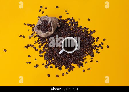 Ein Sack voll Kaffee und ein Stapel Bohnen, die auf einem gelben Hintergrund verstreut sind. In der Mitte aller verstreuten Bohnen ist von einem weißen Porzellan umgeben Stockfoto