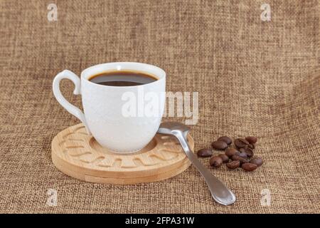 Auf einem Holzteller befindet sich eine weiße Tasse mit Kaffee und einem Metalllöffel. Sie werden auf ein Sacktuch gelegt und es gibt eine Handvoll Kaffeebohnen auf der ri Stockfoto