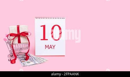 10. Mai. Eine Geschenkbox in einem Einkaufswagen, Dollars und ein Kalender mit dem Datum 10 können auf einem rosa Hintergrund stehen. Frühling Monat, Tag des Jahres Stockfoto