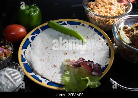 Eine launische Aufnahme zeigt Roti Paratha-Tortilla-Fladenbrot auf Glasplatte, umgeben von frischem Gemüse wie Paprika, Tomaten, Reis und Fleisch Stockfoto