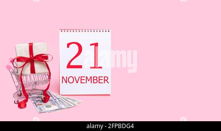 Tag des 21. novembers. Eine Geschenkbox in einem Einkaufswagen, Dollars und ein Kalender mit dem Datum des 21. november auf einem rosa Hintergrund. Herbst Monat, Tag von Stockfoto