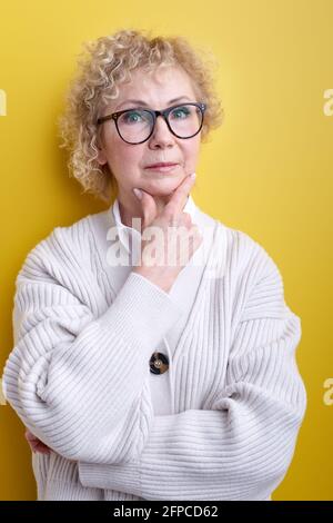 Gealterte Frau mit ernstem und konzentriertem selbstbewussten Blick, Brainstorming und Gedanken an herausfordernde Probleme, posiert isoliert auf Gelb, trägt Eleg Stockfoto