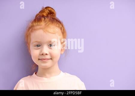 Nettes lächelndes kaukasisches Mädchen mit roten Haaren und großen blauen Augen isoliert über lila Hintergrund. Natürliche rothaarige Mädchen mit Sommersprossen Blick auf Kamer Stockfoto