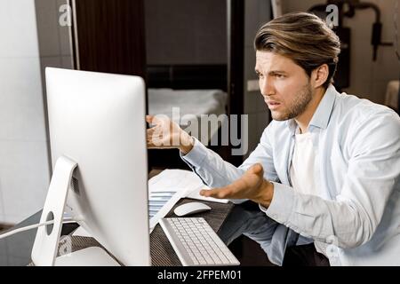 Nachdenkliche männliche Angestellte sitzt am Schreibtisch im Heimbüro, benutzt einen Computer und hat eine Videokonferenz. Verwirrt junger Geschäftsmann im Gespräch mit Stockfoto