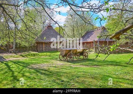 Der alte Holzkarren auf dem grünen Rasen vor den typischen Weidenscheunen mit Strohdächern, Mamajeva Sloboda Kosakendorf, Kiew, Ukraine Stockfoto