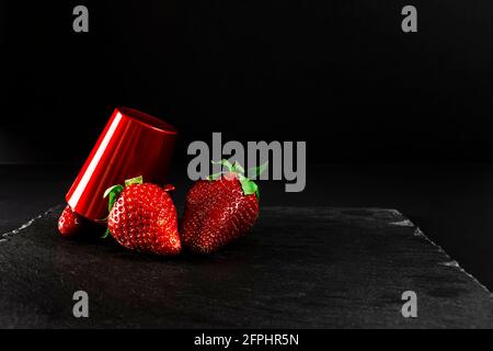Drei frische rote Erdbeeren und eine rote Tasse auf einer Tafel.Es ist ein Stillleben, das komplett mit künstlichem Licht gemacht und im horizontalen Format gedreht wurde. Stockfoto