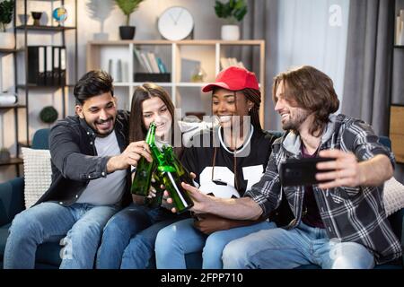 Gruppe von multirassischen Männern und Frauen, die ein modernes Smartphone für Selfie verwenden. Glückliche Freunde sitzen zusammen auf der Couch, trinken Bier und essen Snacks, während sie sich ein Fußballspiel ansehen. Stockfoto