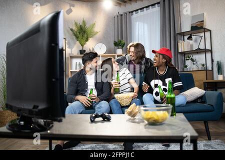 Junge, multiethnische Menschen in legerer Kleidung treffen sich zu Hause, um ein Fußballspiel im Fernsehen zu sehen. Glückliche Freunde unterhalten sich, essen und trinken in der Freizeit. Stockfoto