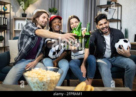 Eine Gruppe von glücklichen multikulturellen Freunden, die zusammen auf der Couch sitzen und Bier trinken und Snacks essen. Fröhliche junge Menschen genießen unbeschwerte Wochenenden zu Hause. Stockfoto