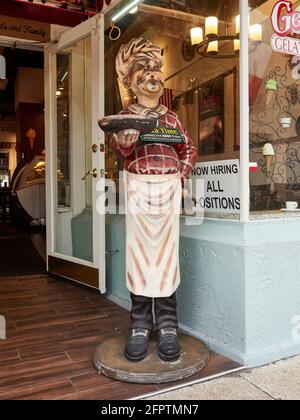 Eine große Statue oder Figur des Küchenchefs vor einem Pizzalestaurant, die eine Pizza mit einem Schild mit Hilfswünschen im Fenster in St. Augustine, Florida, USA, hält. Stockfoto